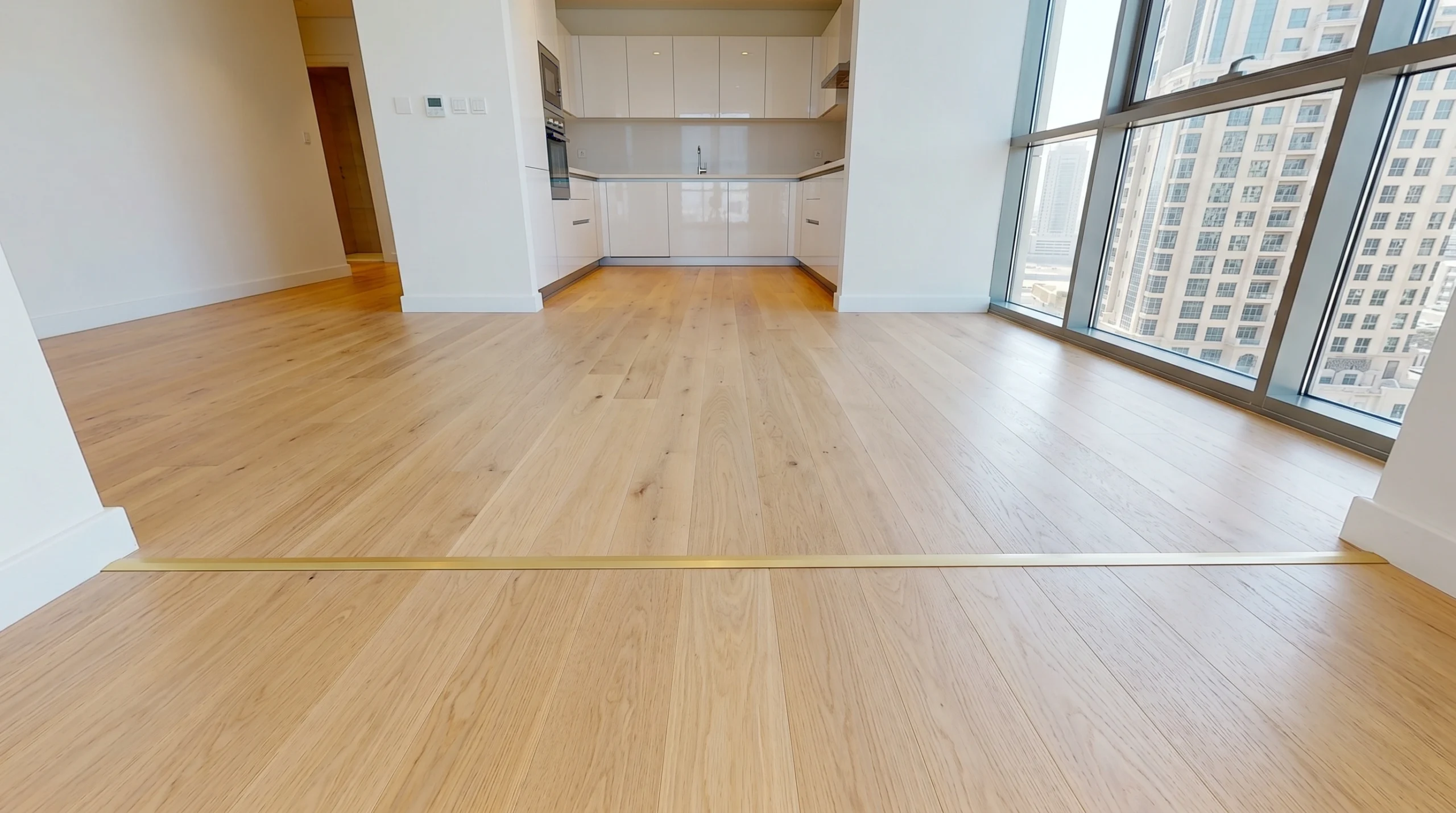 Light ash engineered hardwood flooring flowing continuously from dining area through kitchen threshold in Business Bay Dubai apartment with gold transition strip